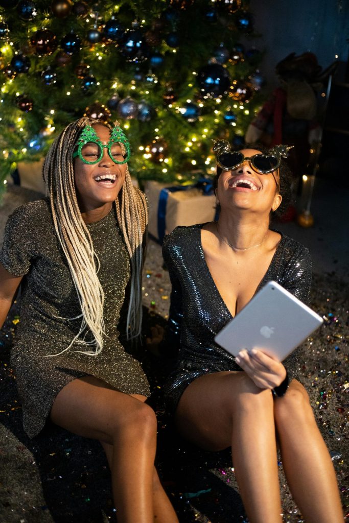 Two women laughing together during a festive Christmas party with fun eyewear and holiday decorations.